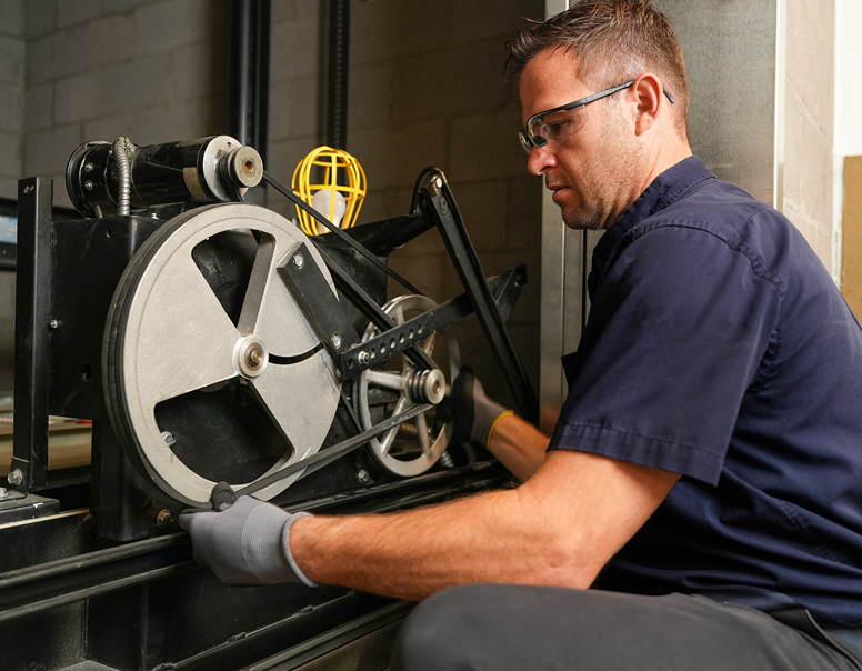Technician in safety glasses working on a belt-driven elevator machine in a machine room